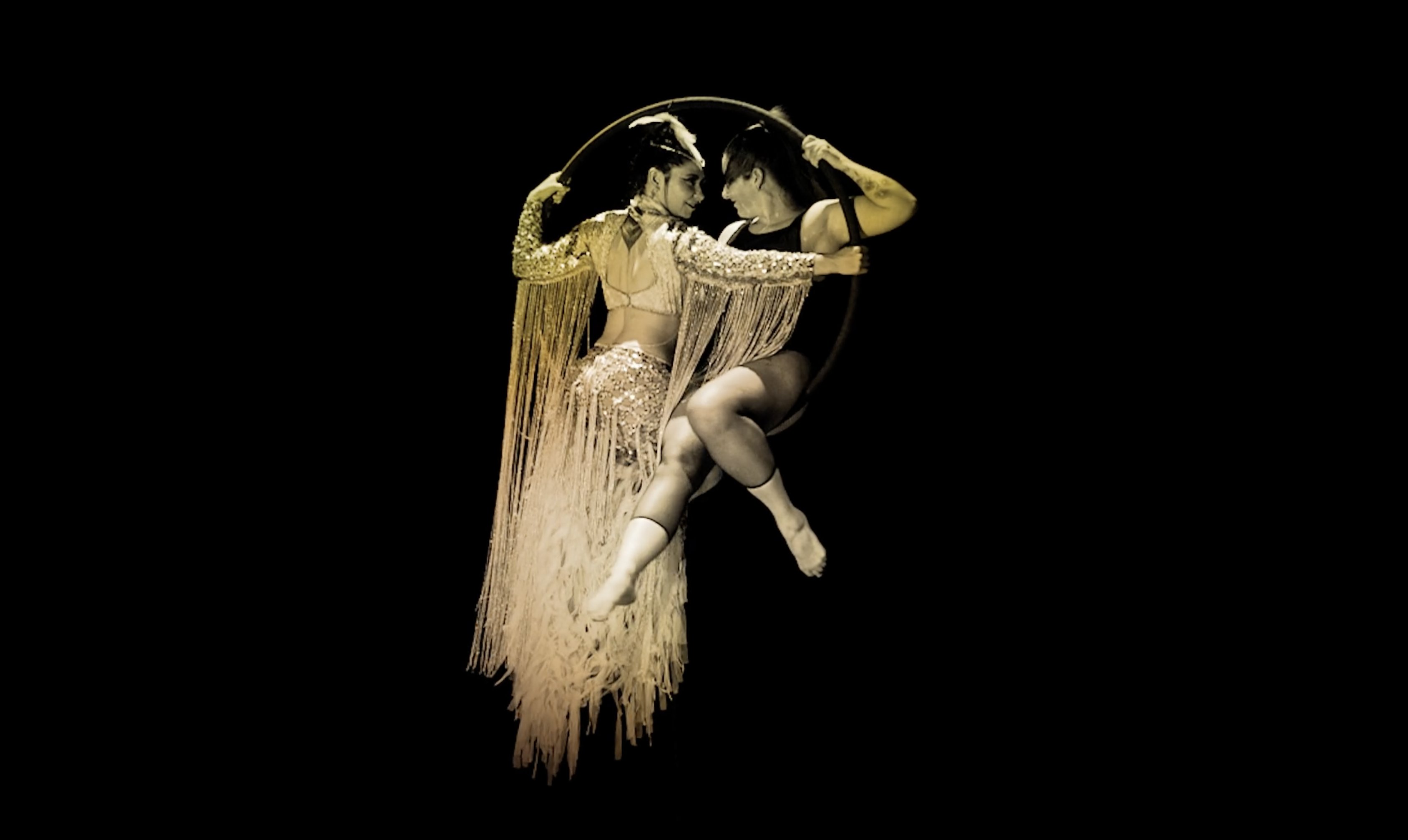 Two aerialists in a hoop, one in a fringed costume, in muted gold tones against a black background.